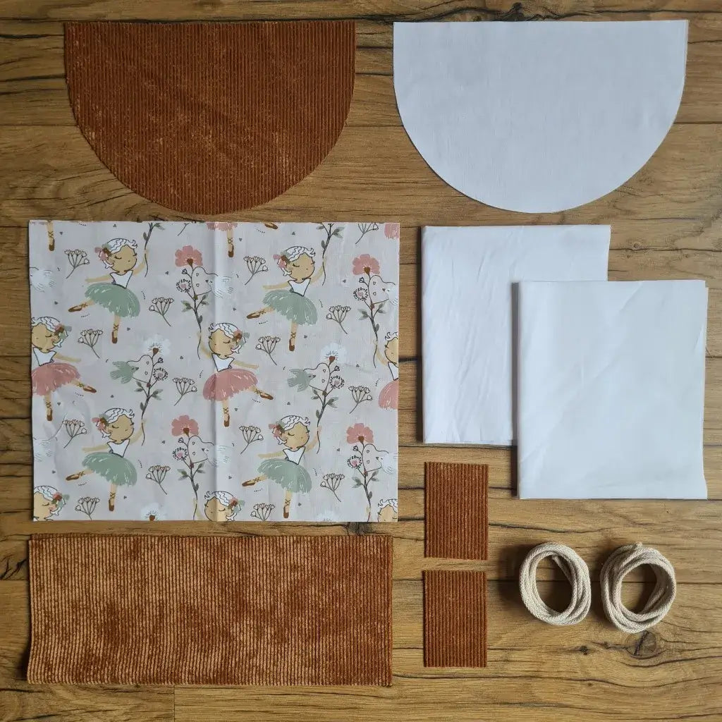 Kit "prêt à coudre" pour un sac à dos format pochon avec rabat pour enfant ou bébé au motif danseuse.  L'ensemble des pièces de tissu, déjà prédécoupées aux bonnes dimensions, sont disposées au sol. Le kit se compose de 2 pièces en coton imprimé au motif danseuse, de 4 pièces en velours côtelé marron, de 3 pièces en tissu blanc pour la doublure du sac et de 2 cordelettes en coton naturel.