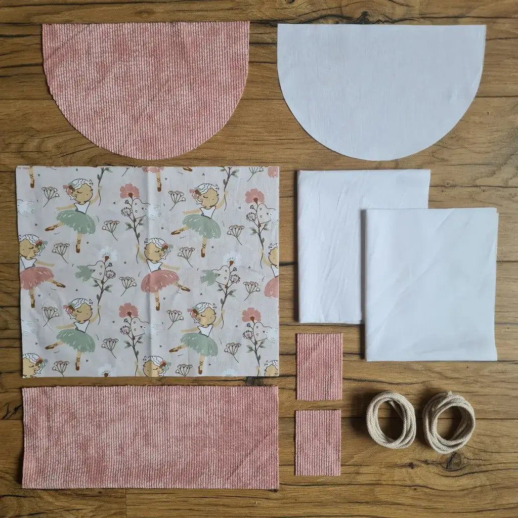 Kit "prêt à coudre" pour un sac à dos format pochon avec rabat pour enfant ou bébé au motif danseuse.  L'ensemble des pièces de tissu, déjà prédécoupées aux bonnes dimensions, sont disposées au sol. Le kit se compose de 2 pièces en coton imprimé au motif danseuse, de 4 pièces en velours côtelé rose, de 3 pièces en tissu blanc pour la doublure du sac et de 2 cordelettes en coton naturel.