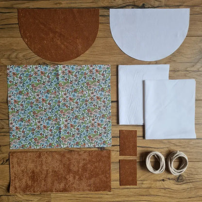 Kit "prêt à coudre" pour un sac à dos format pochon avec rabat pour enfant ou bébé au motif fleurs.  L'ensemble des pièces de tissu, déjà prédécoupées aux bonnes dimensions, sont disposées au sol. Le kit se compose de 2 pièces en coton imprimé au motif fleurs, de 4 pièces en velours côtelé marron, de 3 pièces en tissu blanc pour la doublure du sac et de 2 cordelettes en coton naturel.