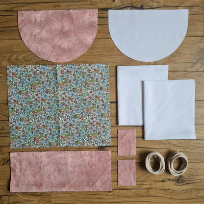 Kit "prêt à coudre" pour un sac à dos format pochon avec rabat pour enfant ou bébé au motif fleurs.  L'ensemble des pièces de tissu, déjà prédécoupées aux bonnes dimensions, sont disposées au sol. Le kit se compose de 2 pièces en coton imprimé au motif fleurs, de 4 pièces en velours côtelé rose, de 3 pièces en tissu blanc pour la doublure du sac et de 2 cordelettes en coton naturel.