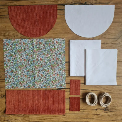 Kit "prêt à coudre" pour un sac à dos format pochon avec rabat pour enfant ou bébé au motif fleurs.  L'ensemble des pièces de tissu, déjà prédécoupées aux bonnes dimensions, sont disposées au sol. Le kit se compose de 2 pièces en coton imprimé au motif fleurs, de 4 pièces en velours côtelé terracotta, de 3 pièces en tissu blanc pour la doublure du sac et de 2 cordelettes en coton naturel.
