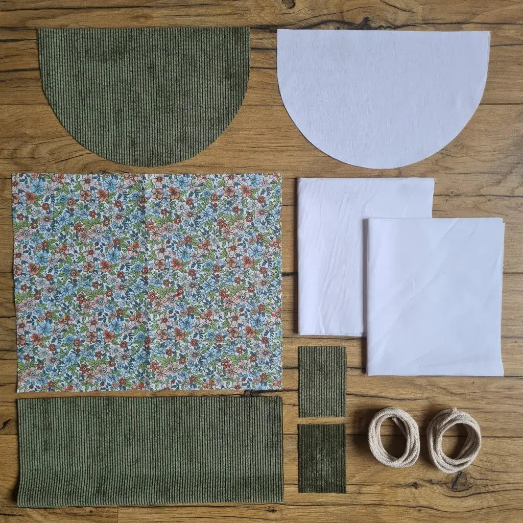 Kit "prêt à coudre" pour un sac à dos format pochon avec rabat pour enfant ou bébé au motif fleurs.  L'ensemble des pièces de tissu, déjà prédécoupées aux bonnes dimensions, sont disposées au sol. Le kit se compose de 2 pièces en coton imprimé au motif fleurs, de 4 pièces en velours côtelé vert, de 3 pièces en tissu blanc pour la doublure du sac et de 2 cordelettes en coton naturel.