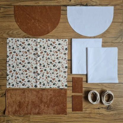 Kit "prêt à coudre" pour un sac à dos format pochon avec rabat pour enfant ou bébé au motif léopard.  L'ensemble des pièces de tissu, déjà prédécoupées aux bonnes dimensions, sont disposées au sol. Le kit se compose de 2 pièces en coton imprimé au motif léopard, de 4 pièces en velours côtelé marron, de 3 pièces en tissu blanc pour la doublure du sac et de 2 cordelettes en coton naturel.