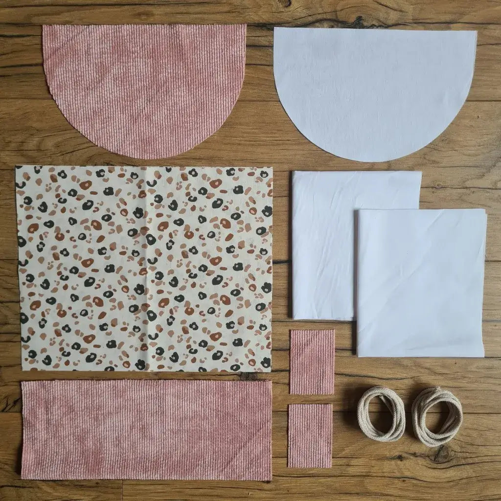 Kit "prêt à coudre" pour un sac à dos format pochon avec rabat pour enfant ou bébé au motif léopard.  L'ensemble des pièces de tissu, déjà prédécoupées aux bonnes dimensions, sont disposées au sol. Le kit se compose de 2 pièces en coton imprimé au motif léopard, de 4 pièces en velours côtelé rose, de 3 pièces en tissu blanc pour la doublure du sac et de 2 cordelettes en coton naturel.