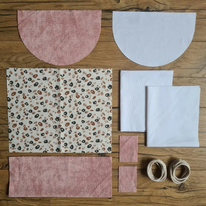 Kit "prêt à coudre" pour un sac à dos format pochon avec rabat pour enfant ou bébé au motif léopard.  L'ensemble des pièces de tissu, déjà prédécoupées aux bonnes dimensions, sont disposées au sol. Le kit se compose de 2 pièces en coton imprimé au motif léopard, de 4 pièces en velours côtelé rose, de 3 pièces en tissu blanc pour la doublure du sac et de 2 cordelettes en coton naturel.