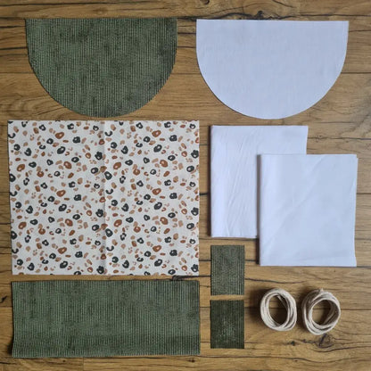Kit "prêt à coudre" pour un sac à dos format pochon avec rabat pour enfant ou bébé au motif léopard.  L'ensemble des pièces de tissu, déjà prédécoupées aux bonnes dimensions, sont disposées au sol. Le kit se compose de 2 pièces en coton imprimé au motif léopard, de 4 pièces en velours côtelé vert, de 3 pièces en tissu blanc pour la doublure du sac et de 2 cordelettes en coton naturel.