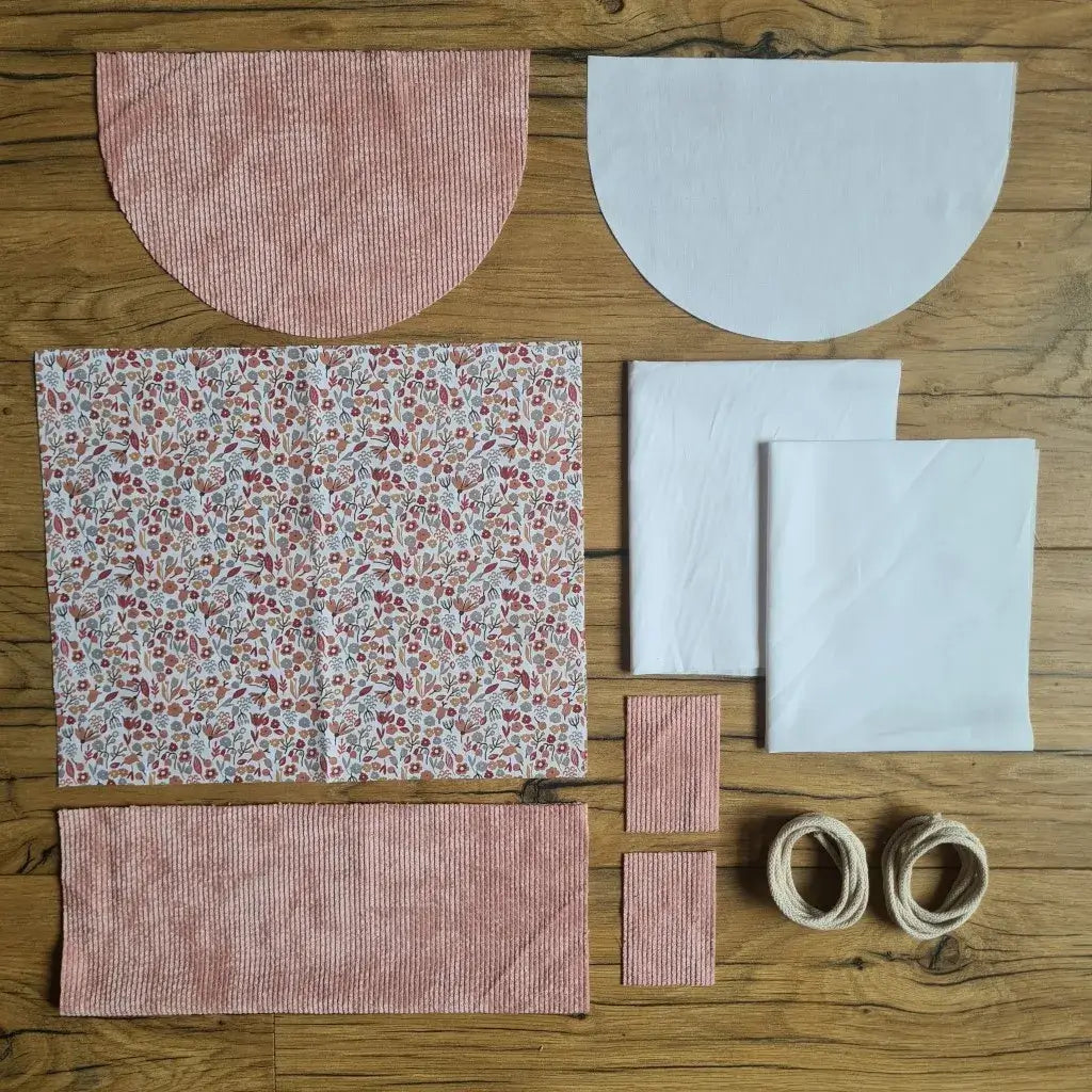 Kit "prêt à coudre" pour un sac à dos format pochon avec rabat pour enfant ou bébé au motif liberty.  L'ensemble des pièces de tissu, déjà prédécoupées aux bonnes dimensions, sont disposées au sol. Le kit se compose de 2 pièces en coton imprimé au motif liberty, de 4 pièces en velours côtelé rose, de 3 pièces en tissu blanc pour la doublure du sac et de 2 cordelettes en coton naturel.