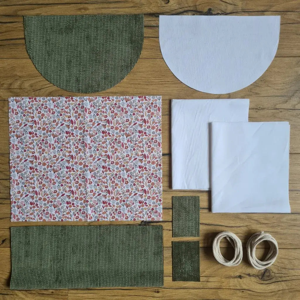 Kit "prêt à coudre" pour un sac à dos format pochon avec rabat pour enfant ou bébé au motif liberty.  L'ensemble des pièces de tissu, déjà prédécoupées aux bonnes dimensions, sont disposées au sol. Le kit se compose de 2 pièces en coton imprimé au motif liberty, de 4 pièces en velours côtelé vert, de 3 pièces en tissu blanc pour la doublure du sac et de 2 cordelettes en coton naturel.