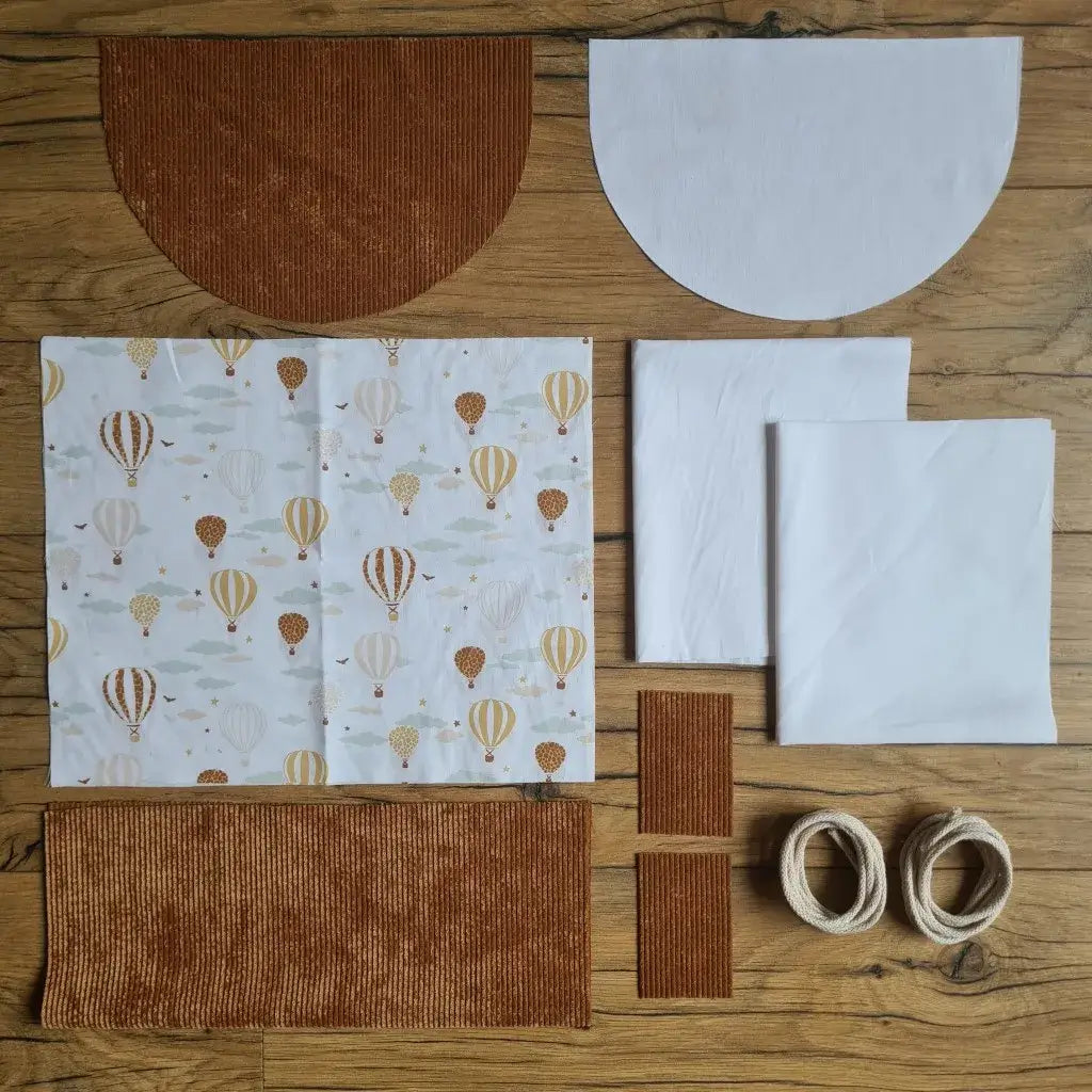 Kit "prêt à coudre" pour un sac à dos format pochon avec rabat pour enfant ou bébé au motif montgolfières.  L'ensemble des pièces de tissu, déjà prédécoupées aux bonnes dimensions, sont disposées au sol. Le kit se compose de 2 pièces en coton imprimé au motif montgolfières, de 4 pièces en velours côtelé marron, de 3 pièces en tissu blanc pour la doublure du sac et de 2 cordelettes en coton naturel.