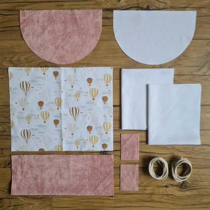 Kit "prêt à coudre" pour un sac à dos format pochon avec rabat pour enfant ou bébé au motif montgolfières.  L'ensemble des pièces de tissu, déjà prédécoupées aux bonnes dimensions, sont disposées au sol. Le kit se compose de 2 pièces en coton imprimé au motif montgolfières, de 4 pièces en velours côtelé rose, de 3 pièces en tissu blanc pour la doublure du sac et de 2 cordelettes en coton naturel.
