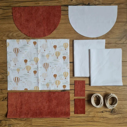 Kit "prêt à coudre" pour un sac à dos format pochon avec rabat pour enfant ou bébé au motif montgolfières.  L'ensemble des pièces de tissu, déjà prédécoupées aux bonnes dimensions, sont disposées au sol. Le kit se compose de 2 pièces en coton imprimé au motif montgolfières, de 4 pièces en velours côtelé terracotta, de 3 pièces en tissu blanc pour la doublure du sac et de 2 cordelettes en coton naturel.