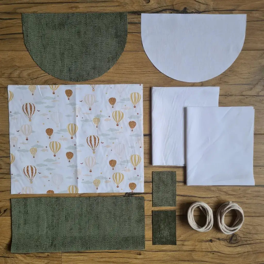 Kit "prêt à coudre" pour un sac à dos format pochon avec rabat pour enfant ou bébé au motif montgolfières.  L'ensemble des pièces de tissu, déjà prédécoupées aux bonnes dimensions, sont disposées au sol. Le kit se compose de 2 pièces en coton imprimé au motif montgolfières, de 4 pièces en velours côtelé vert, de 3 pièces en tissu blanc pour la doublure du sac et de 2 cordelettes en coton naturel.