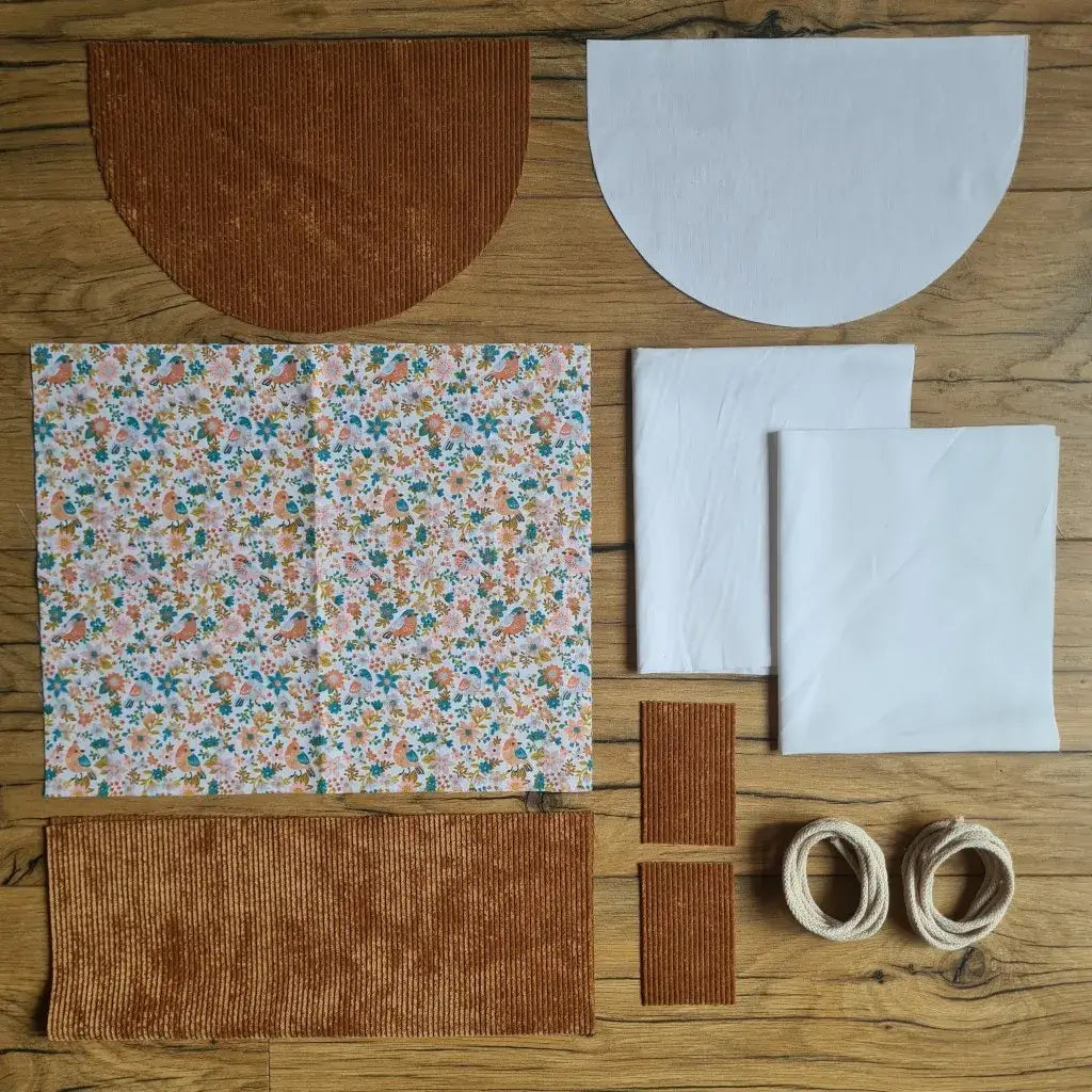 Kit "prêt à coudre" pour un sac à dos format pochon avec rabat pour enfant ou bébé au motif oiseaux et fleurs.  L'ensemble des pièces de tissu, déjà prédécoupées aux bonnes dimensions, sont disposées au sol. Le kit se compose de 2 pièces en coton imprimé au motif oiseaux et fleurs, de 4 pièces en velours côtelé marron, de 3 pièces en tissu blanc pour la doublure du sac et de 2 cordelettes en coton naturel.