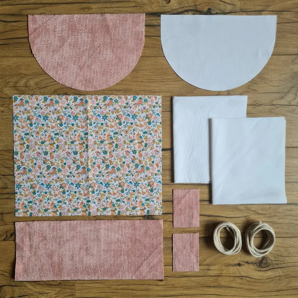 Kit "prêt à coudre" pour un sac à dos format pochon avec rabat pour enfant ou bébé au motif oiseaux et fleurs.  L'ensemble des pièces de tissu, déjà prédécoupées aux bonnes dimensions, sont disposées au sol. Le kit se compose de 2 pièces en coton imprimé au motif oiseaux et fleurs, de 4 pièces en velours côtelé rose, de 3 pièces en tissu blanc pour la doublure du sac et de 2 cordelettes en coton naturel.