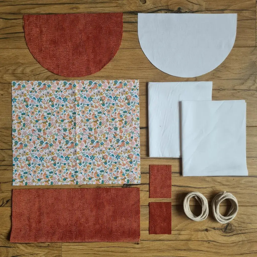 Kit "prêt à coudre" pour un sac à dos format pochon avec rabat pour enfant ou bébé au motif oiseaux et fleurs.  L'ensemble des pièces de tissu, déjà prédécoupées aux bonnes dimensions, sont disposées au sol. Le kit se compose de 2 pièces en coton imprimé au motif oiseaux et fleurs, de 4 pièces en velours côtelé terracotta, de 3 pièces en tissu blanc pour la doublure du sac et de 2 cordelettes en coton naturel.