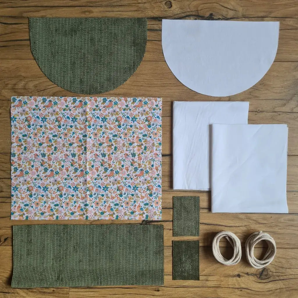 Kit "prêt à coudre" pour un sac à dos format pochon avec rabat pour enfant ou bébé au motif oiseaux et fleurs.  L'ensemble des pièces de tissu, déjà prédécoupées aux bonnes dimensions, sont disposées au sol. Le kit se compose de 2 pièces en coton imprimé au motif oiseaux et fleurs, de 4 pièces en velours côtelé vert, de 3 pièces en tissu blanc pour la doublure du sac et de 2 cordelettes en coton naturel.