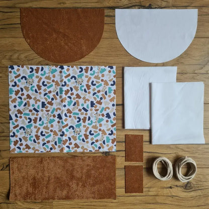 Kit "prêt à coudre" pour un sac à dos format pochon avec rabat pour enfant ou bébé au motif ptiwi.  L'ensemble des pièces de tissu, déjà prédécoupées aux bonnes dimensions, sont disposées au sol. Le kit se compose de 2 pièces en coton imprimé au motif ptiwi, de 4 pièces en velours côtelé marron, de 3 pièces en tissu blanc pour la doublure du sac et de 2 cordelettes en coton naturel.