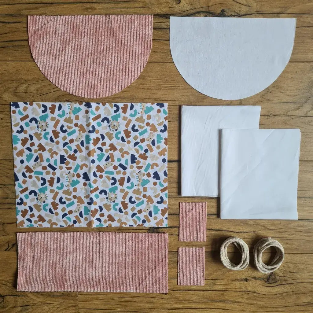 Kit "prêt à coudre" pour un sac à dos format pochon avec rabat pour enfant ou bébé au motif ptiwi.  L'ensemble des pièces de tissu, déjà prédécoupées aux bonnes dimensions, sont disposées au sol. Le kit se compose de 2 pièces en coton imprimé au motif ptiwi, de 4 pièces en velours côtelé rose, de 3 pièces en tissu blanc pour la doublure du sac et de 2 cordelettes en coton naturel.