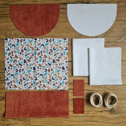 Kit "prêt à coudre" pour un sac à dos format pochon avec rabat pour enfant ou bébé au motif ptiwi.  L'ensemble des pièces de tissu, déjà prédécoupées aux bonnes dimensions, sont disposées au sol. Le kit se compose de 2 pièces en coton imprimé au motif ptiwi, de 4 pièces en velours côtelé terracotta, de 3 pièces en tissu blanc pour la doublure du sac et de 2 cordelettes en coton naturel.