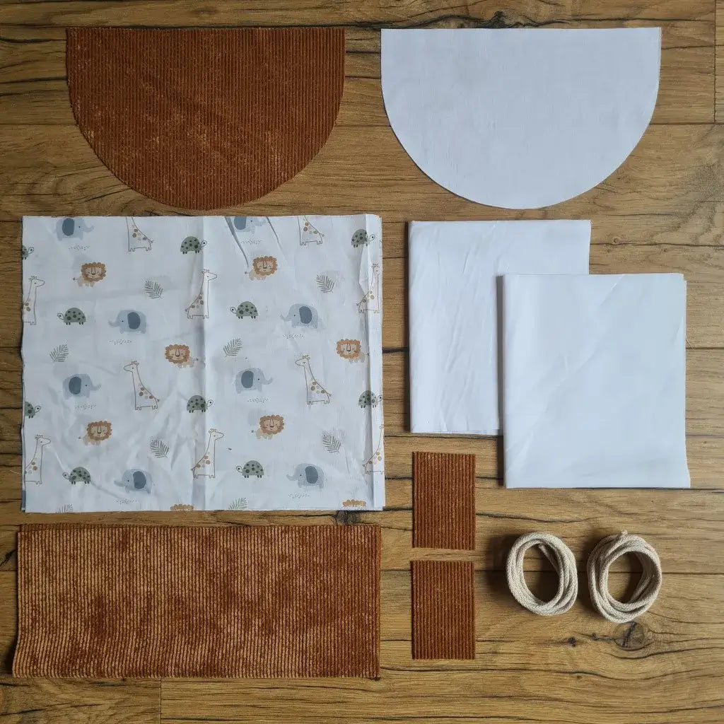 Kit "prêt à coudre" pour un sac à dos format pochon avec rabat pour enfant ou bébé au motif savane.  L'ensemble des pièces de tissu, déjà prédécoupées aux bonnes dimensions, sont disposées au sol. Le kit se compose de 2 pièces en coton imprimé au motif savane, de 4 pièces en velours côtelé marron, de 3 pièces en tissu blanc pour la doublure du sac et de 2 cordelettes en coton naturel.