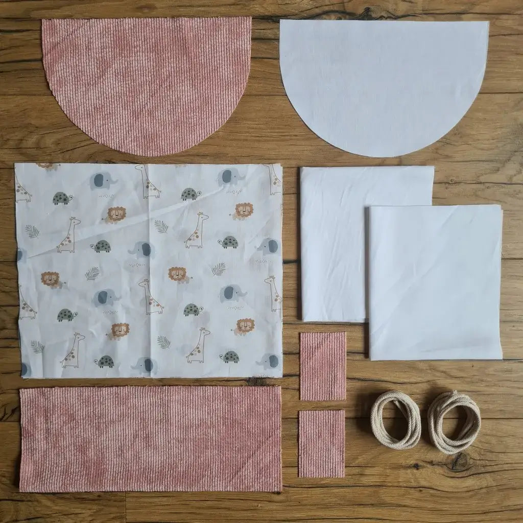 Kit "prêt à coudre" pour un sac à dos format pochon avec rabat pour enfant ou bébé au motif savane.  L'ensemble des pièces de tissu, déjà prédécoupées aux bonnes dimensions, sont disposées au sol. Le kit se compose de 2 pièces en coton imprimé au motif savane, de 4 pièces en velours côtelé rose, de 3 pièces en tissu blanc pour la doublure du sac et de 2 cordelettes en coton naturel.