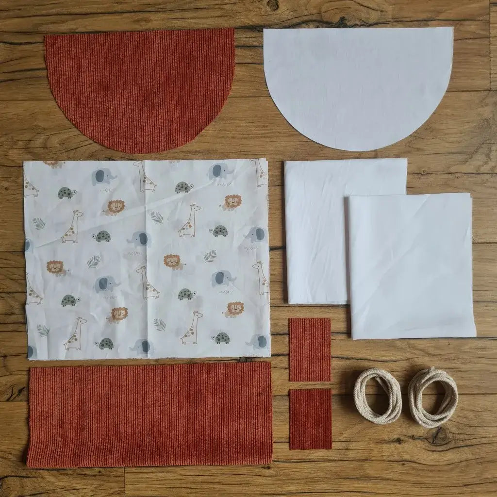 Kit "prêt à coudre" pour un sac à dos format pochon avec rabat pour enfant ou bébé au motif savane.  L'ensemble des pièces de tissu, déjà prédécoupées aux bonnes dimensions, sont disposées au sol. Le kit se compose de 2 pièces en coton imprimé au motif savane, de 4 pièces en velours côtelé terracotta, de 3 pièces en tissu blanc pour la doublure du sac et de 2 cordelettes en coton naturel.