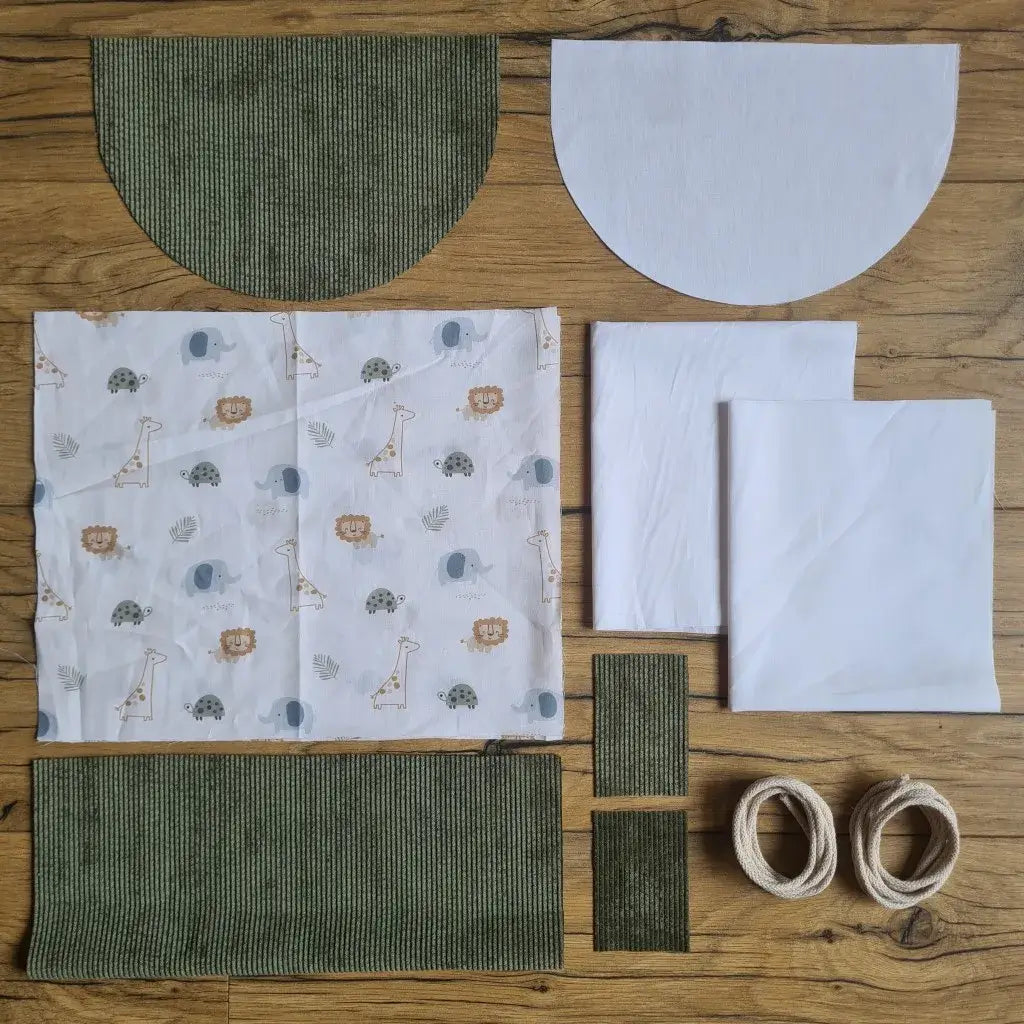 Kit "prêt à coudre" pour un sac à dos format pochon avec rabat pour enfant ou bébé au motif savane.  L'ensemble des pièces de tissu, déjà prédécoupées aux bonnes dimensions, sont disposées au sol. Le kit se compose de 2 pièces en coton imprimé au motif savane, de 4 pièces en velours côtelé vert, de 3 pièces en tissu blanc pour la doublure du sac et de 2 cordelettes en coton naturel.