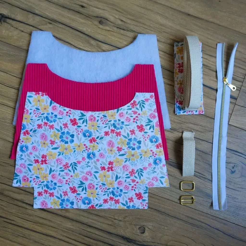 Kit prêt-à-coudre pour sac bandoulière, comprenant des pièces de tissu découpées en velours côtelé framboise et en coton imprimé floral multicolore aux tons jaune, rose, bleu et vert. Sont également inclus : une fermeture éclair blanche à curseur doré, une sangle en coton beige, de la ouate de renfort et la bouclerie dorée pour le réglage de la sangle. Présenté sur un sol en bois, ce kit permet de réaliser facilement un sac artisanal unique et personnalisé.