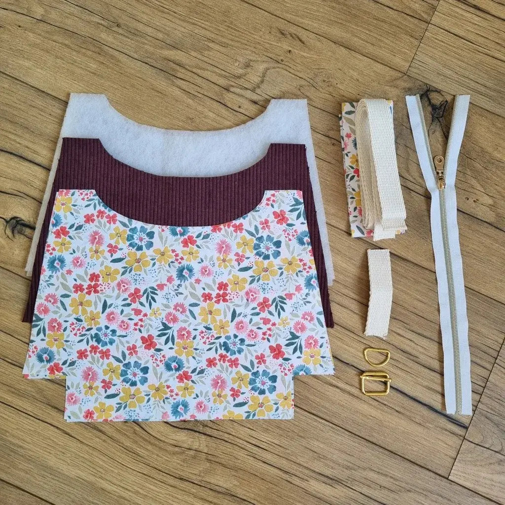 Kit couture complet pour réaliser un sac bandoulière en velours côtelé prune et coton fleuri multicolore, comprenant les tissus découpés, la sangle beige, la fermeture éclair dorée, les boucles métalliques et les pièces de ouate. Projet DIY idéal pour couturières débutantes souhaitant créer un sac pratique et élégant fait main.