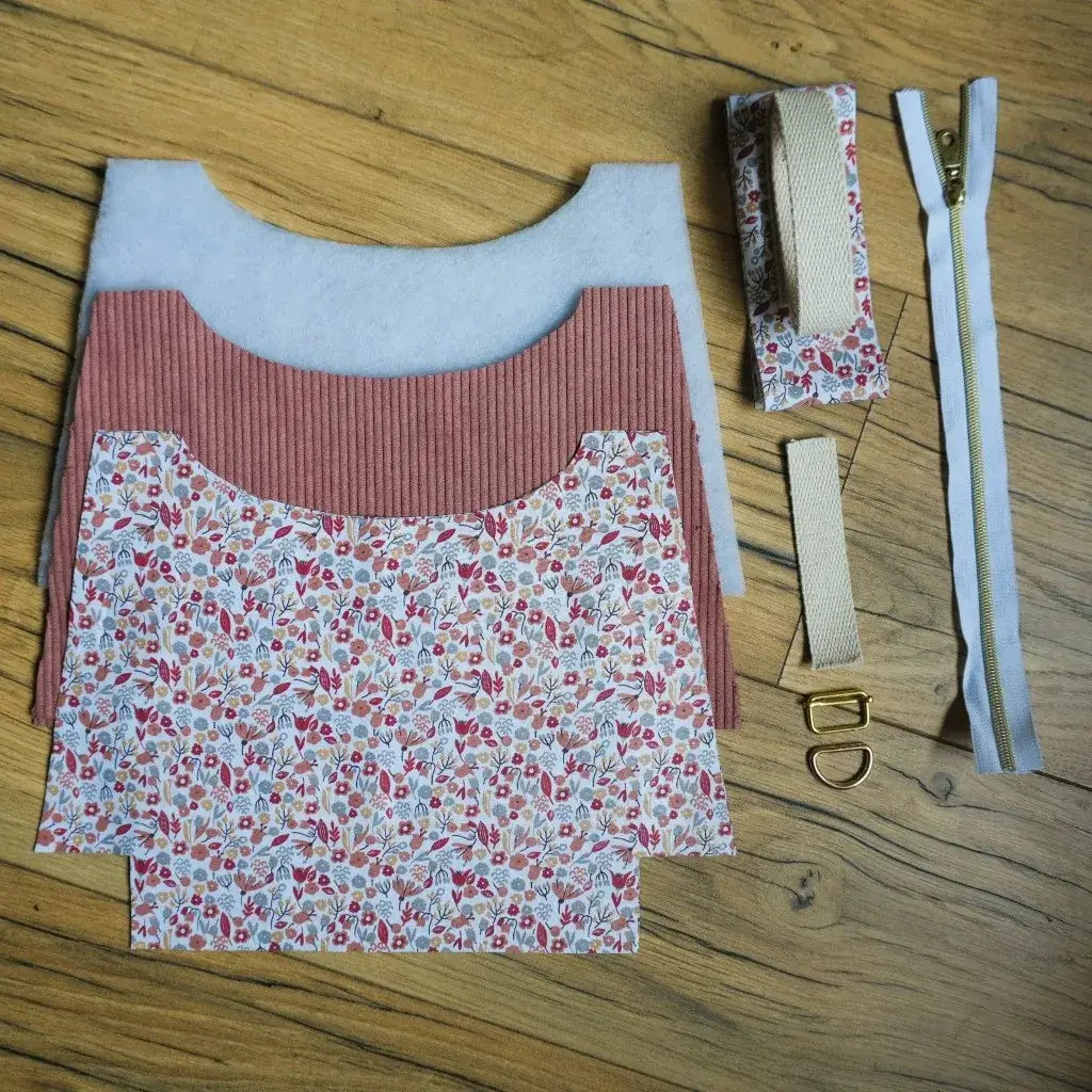 Kit prêt-à-coudre pour sac bandoulière composé de pièces de tissu en velours côtelé rose poudré et d’un coton imprimé fleuri aux tons rouges, roses et verts. Le lot inclut également une fermeture éclair blanche à curseur doré, une sangle en coton beige, de la ouate pour la structure et des accessoires métalliques dorés (anneau et boucle). Présenté sur un sol en bois, ce kit permet de créer facilement un sac artisanal unique et élégant.
