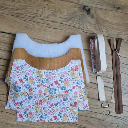 Kit prêt-à-coudre pour sac bandoulière en velours côtelé jaune moutarde, comprenant les pièces de tissu extérieur, doublure fleurie multicolore, sangle coton, fermeture éclair marron, boucles en métal et bande assortie. Le tout est disposé sur un sol en bois naturel. Idéal pour confectionner soi-même un sac fait main au style bohème, coloré et tendance, accessible même aux débutantes.