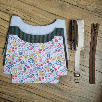 Kit prêt à coudre pour sac bandoulière composé de pièces de tissu en velours côtelé vert, coton fleuri, renfort thermocollant, sangle en coton, zip marron et boucles métalliques, le tout posé sur un sol en bois. Ce kit DIY complet permet de créer un sac élégant et personnalisé, parfait pour les amateurs de couture créative.