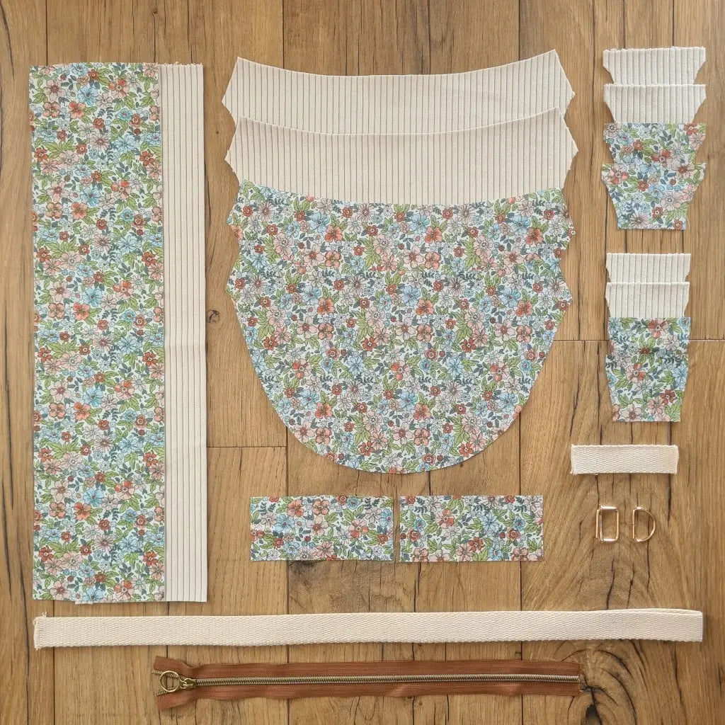 Kit "prêt à coudre" pour un sac demi-lune en velours côtelé au coloris beige avec une doublure en tissu à motif fleuri. L'ensemble des pièces de tissu, déjà prédécoupées aux bonnes dimensions, sont disposées au sol et comprennent neuf pièces en tissu à motif fleuri et sept pièces en velours côtelé de couleur beige ainsi qu'une fermeture éclair marron et une sangle beige.