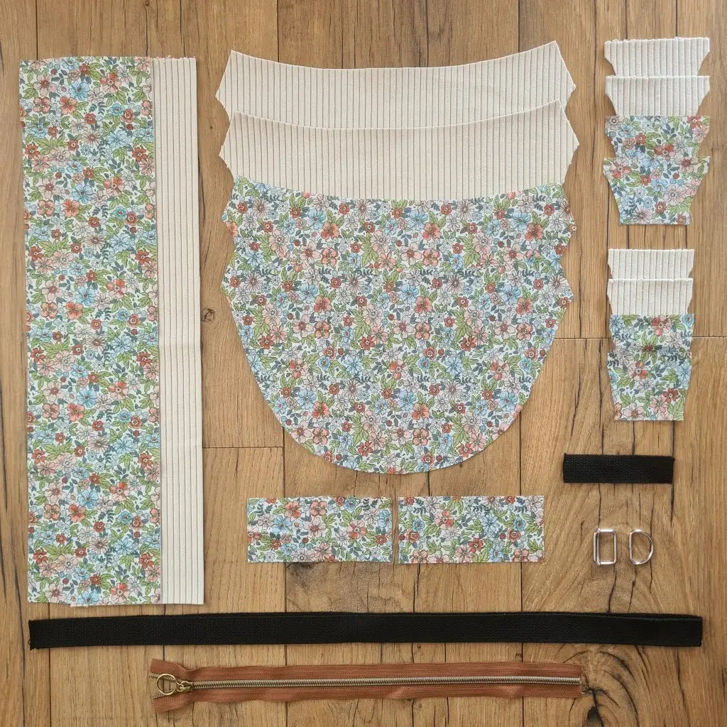 Kit "prêt à coudre" pour un sac demi-lune en velours côtelé au coloris beige avec une doublure en tissu à motif fleuri. L'ensemble des pièces de tissu, déjà prédécoupées aux bonnes dimensions, sont disposées au sol et comprennent neuf pièces en tissu à motif fleuri et sept pièces en velours côtelé de couleur beige ainsi qu'une fermeture éclair marron et une sangle noire.