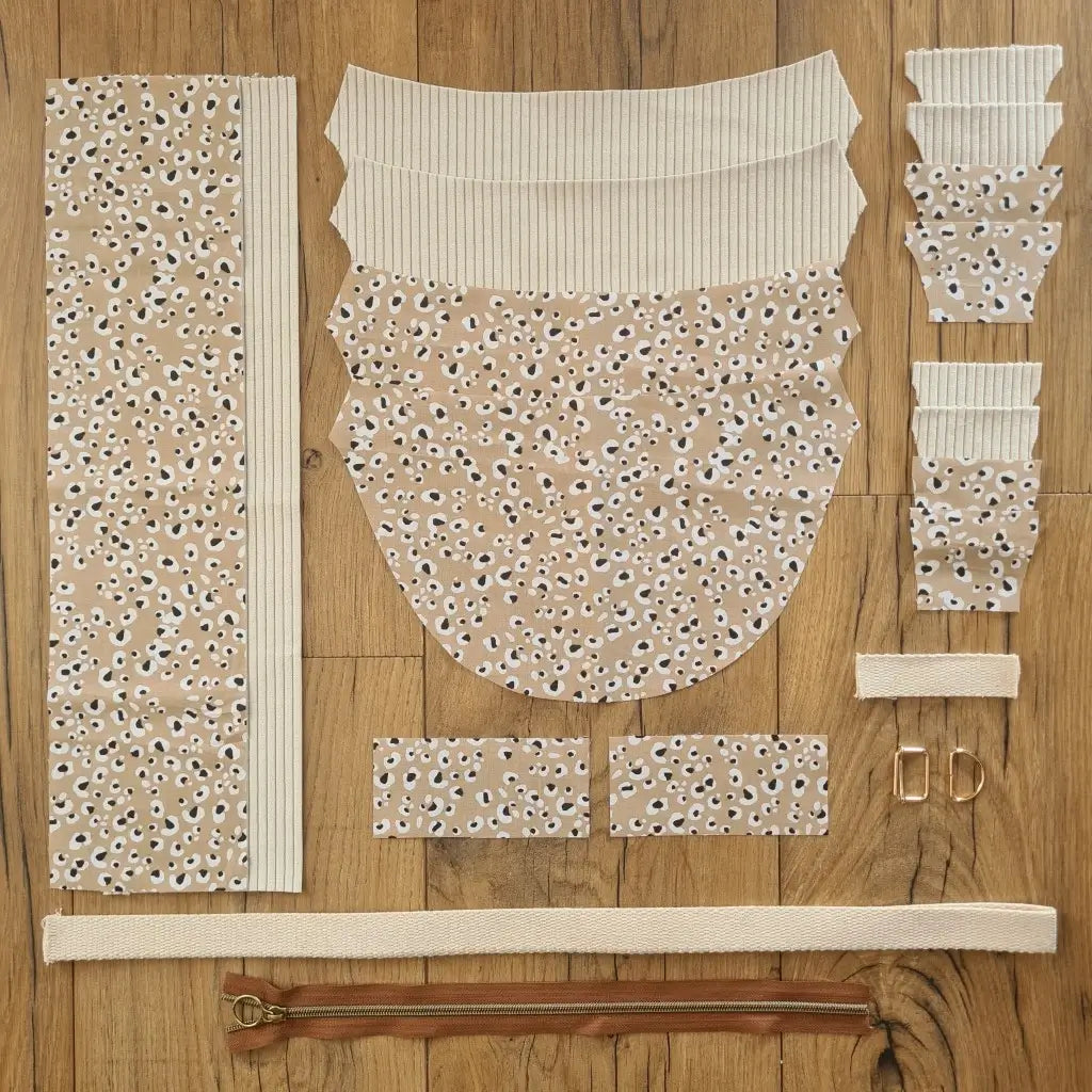 Kit "prêt à coudre" pour un sac demi-lune en velours côtelé au coloris beige avec une doublure en tissu à motif léopard. L'ensemble des pièces de tissu, déjà prédécoupées aux bonnes dimensions, sont disposées au sol et comprennent neuf pièces en tissu à motif léopard et sept pièces en velours côtelé de couleur beige ainsi qu'une fermeture éclair marron et une sangle beige.