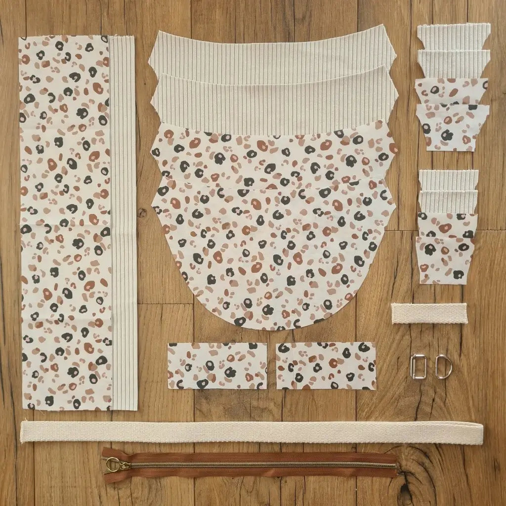 Kit "prêt à coudre" pour un sac demi-lune en velours côtelé au coloris beige avec une doublure en tissu à motif léopard. L'ensemble des pièces de tissu, déjà prédécoupées aux bonnes dimensions, sont disposées au sol et comprennent neuf pièces en tissu à motif léopard et sept pièces en velours côtelé de couleur beige ainsi qu'une fermeture éclair marron et une sangle beige.