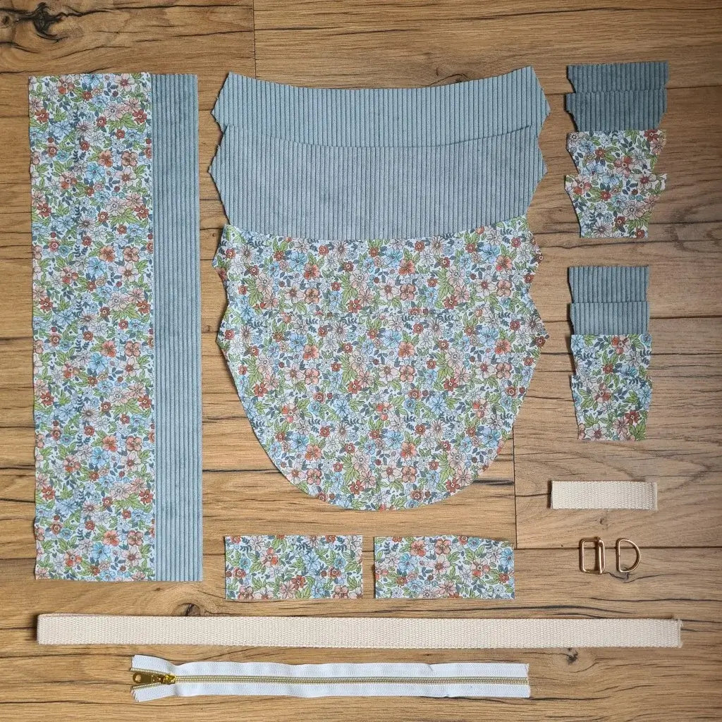 Kit "prêt à coudre" pour un sac demi-lune en velours côtelé au coloris bleu ciel avec une doublure en tissu à motif fleuri. L'ensemble des pièces de tissu, déjà prédécoupées aux bonnes dimensions, sont disposées au sol et comprennent neuf pièces en tissu à motif fleuri et sept pièces en velours côtelé de couleur bleu ciel ainsi qu'une fermeture éclair blanche et une sangle beige.