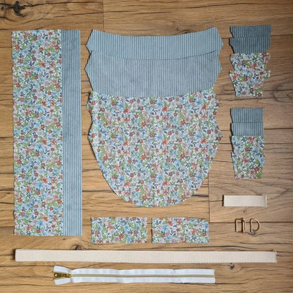 Kit "prêt à coudre" pour un sac demi-lune en velours côtelé au coloris bleu ciel avec une doublure en tissu à motif fleuri. L'ensemble des pièces de tissu, déjà prédécoupées aux bonnes dimensions, sont disposées au sol et comprennent neuf pièces en tissu à motif fleuri et sept pièces en velours côtelé de couleur bleu ciel ainsi qu'une fermeture éclair blanche et une sangle beige.