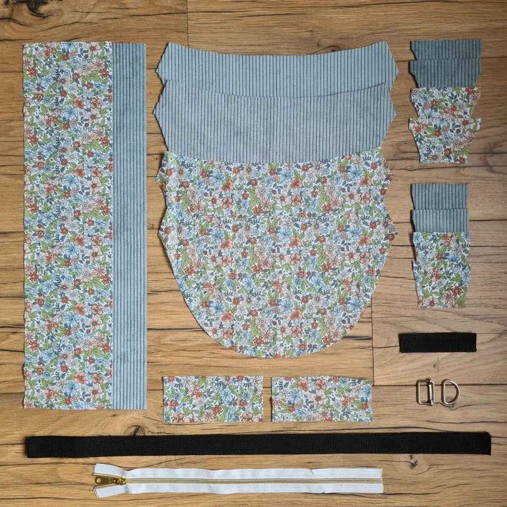 Kit "prêt à coudre" pour un sac demi-lune en velours côtelé au coloris bleu ciel avec une doublure en tissu à motif fleuri. L'ensemble des pièces de tissu, déjà prédécoupées aux bonnes dimensions, sont disposées au sol et comprennent neuf pièces en tissu à motif fleuri et sept pièces en velours côtelé de couleur bleu ciel ainsi qu'une fermeture éclair blanche et une sangle noire.