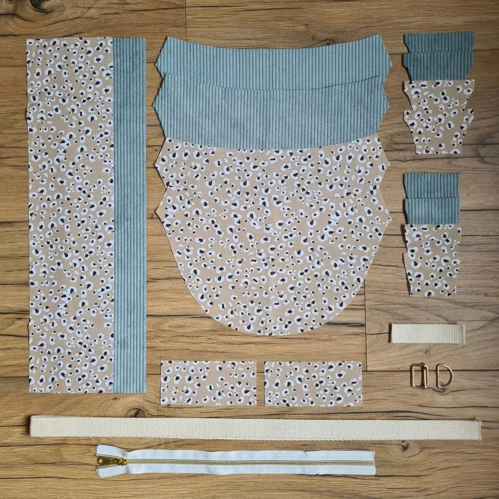 Kit "prêt à coudre" pour un sac demi-lune en velours côtelé au coloris bleu ciel avec une doublure en tissu à motif léopard. L'ensemble des pièces de tissu, déjà prédécoupées aux bonnes dimensions, sont disposées au sol et comprennent neuf pièces en tissu à motif léopard et sept pièces en velours côtelé de couleur bleu ciel ainsi qu'une fermeture éclair blanche et une sangle beige.