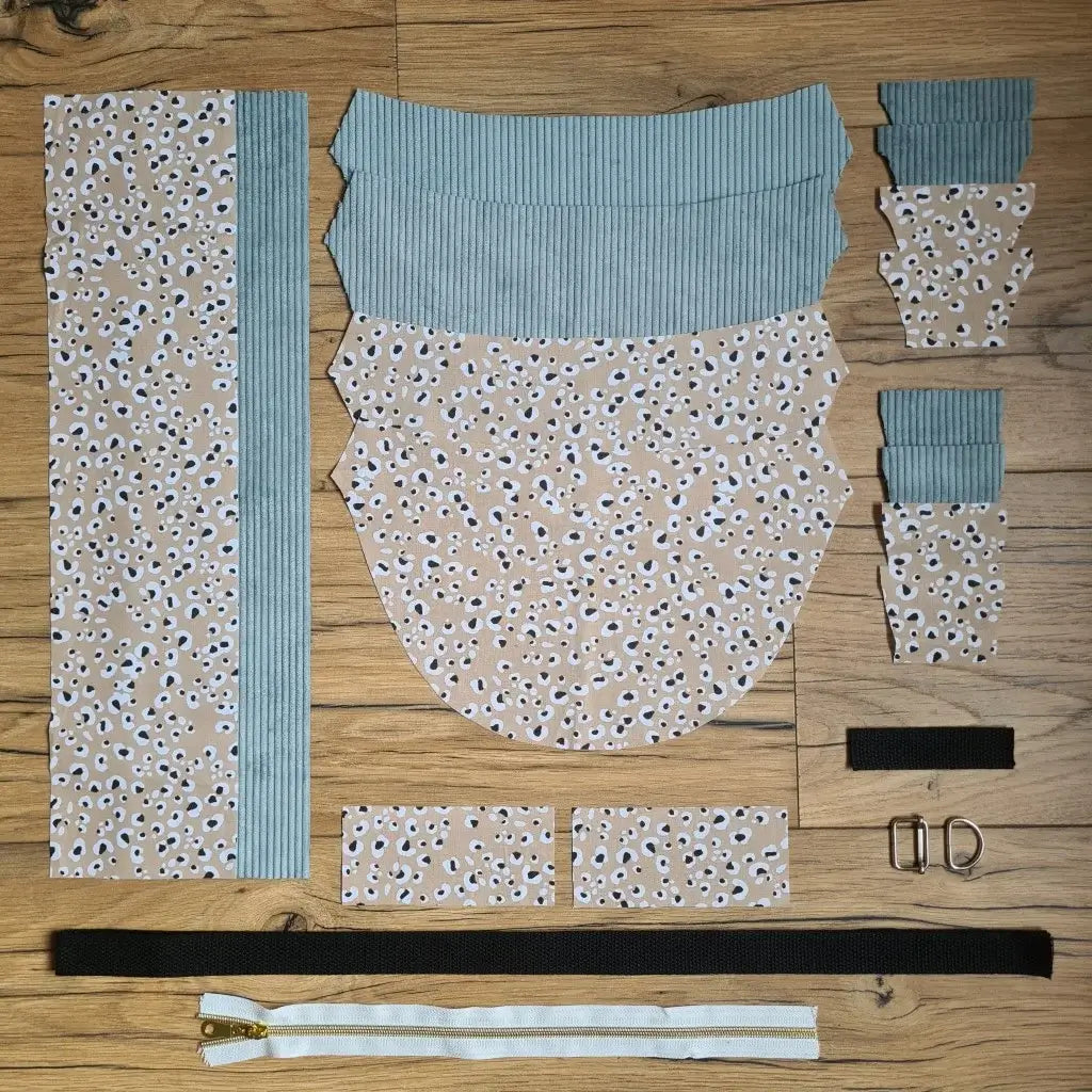 Kit "prêt à coudre" pour un sac demi-lune en velours côtelé au coloris bleu ciel avec une doublure en tissu à motif léopard. L'ensemble des pièces de tissu, déjà prédécoupées aux bonnes dimensions, sont disposées au sol et comprennent neuf pièces en tissu à motif léopard et sept pièces en velours côtelé de couleur bleu ciel ainsi qu'une fermeture éclair blanche et une sangle noire.