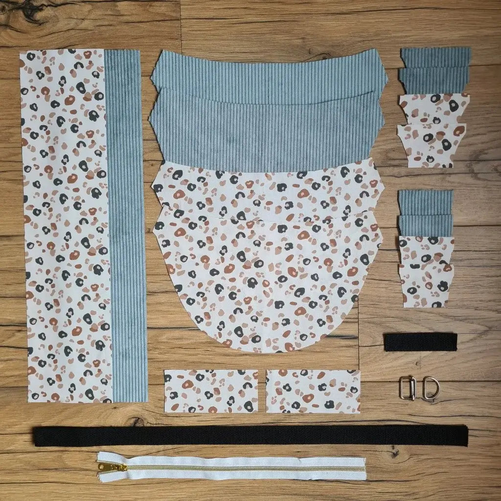 Kit "prêt à coudre" pour un sac demi-lune en velours côtelé au coloris bleu ciel avec une doublure en tissu à motif léopard. L'ensemble des pièces de tissu, déjà prédécoupées aux bonnes dimensions, sont disposées au sol et comprennent neuf pièces en tissu à motif léopard et sept pièces en velours côtelé de couleur bleu ciel ainsi qu'une fermeture éclair blanche et une sangle noire.