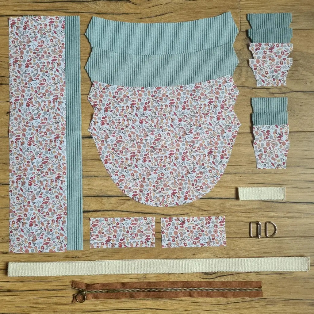 Kit "prêt à coudre" pour un sac demi-lune en velours côtelé au coloris bleu clair avec une doublure en tissu à motif liberty. L'ensemble des pièces de tissu, déjà prédécoupées aux bonnes dimensions, sont disposées au sol et comprennent neuf pièces en tissu à motif liberty et sept pièces en velours côtelé de couleur bleu clair ainsi qu'une fermeture éclair marron et une sangle beige.