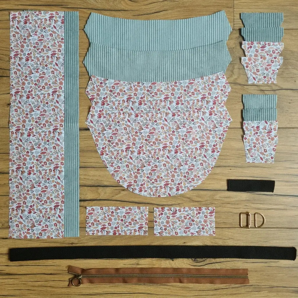 Kit "prêt à coudre" pour un sac demi-lune en velours côtelé au coloris bleu clair avec une doublure en tissu à motif liberty. L'ensemble des pièces de tissu, déjà prédécoupées aux bonnes dimensions, sont disposées au sol et comprennent neuf pièces en tissu à motif liberty et sept pièces en velours côtelé de couleur bleu clair ainsi qu'une fermeture éclair marron et une sangle noire.