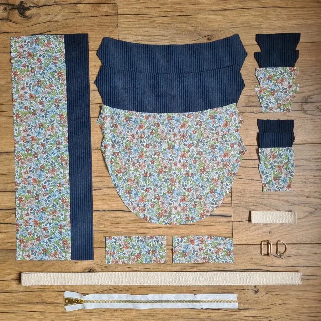 Kit "prêt à coudre" pour un sac demi-lune en velours côtelé au coloris bleu foncé avec une doublure en tissu à motif fleuri. L'ensemble des pièces de tissu, déjà prédécoupées aux bonnes dimensions, sont disposées au sol et comprennent neuf pièces en tissu à motif fleuri et sept pièces en velours côtelé de couleur bleu foncé ainsi qu'une fermeture éclair blanche et une sangle beige.