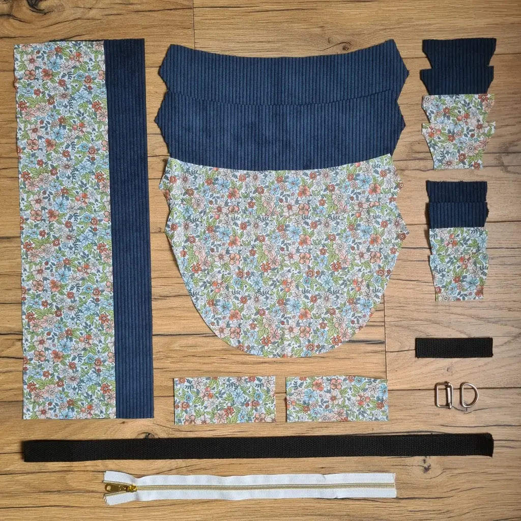 Kit "prêt à coudre" pour un sac demi-lune en velours côtelé au coloris bleu foncé avec une doublure en tissu à motif fleuri. L'ensemble des pièces de tissu, déjà prédécoupées aux bonnes dimensions, sont disposées au sol et comprennent neuf pièces en tissu à motif fleuri et sept pièces en velours côtelé de couleur bleu foncé ainsi qu'une fermeture éclair blanche et une sangle noire.