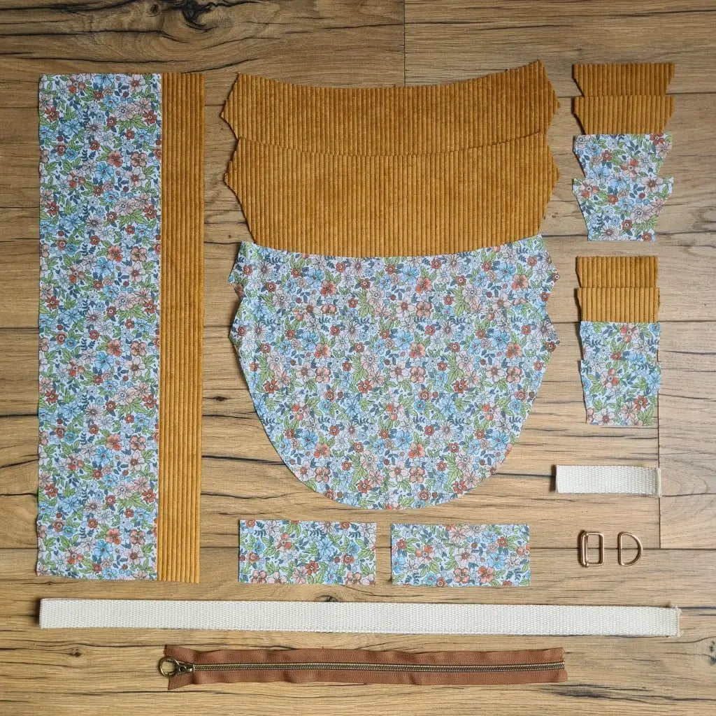 Kit "prêt à coudre" pour un sac demi-lune en velours côtelé au coloris jaune avec une doublure en tissu à motif fleuri. L'ensemble des pièces de tissu, déjà prédécoupées aux bonnes dimensions, sont disposées au sol et comprennent neuf pièces en tissu à motif fleuri et sept pièces en velours côtelé de couleur jaune ainsi qu'une fermeture éclair marron et une sangle blanche.