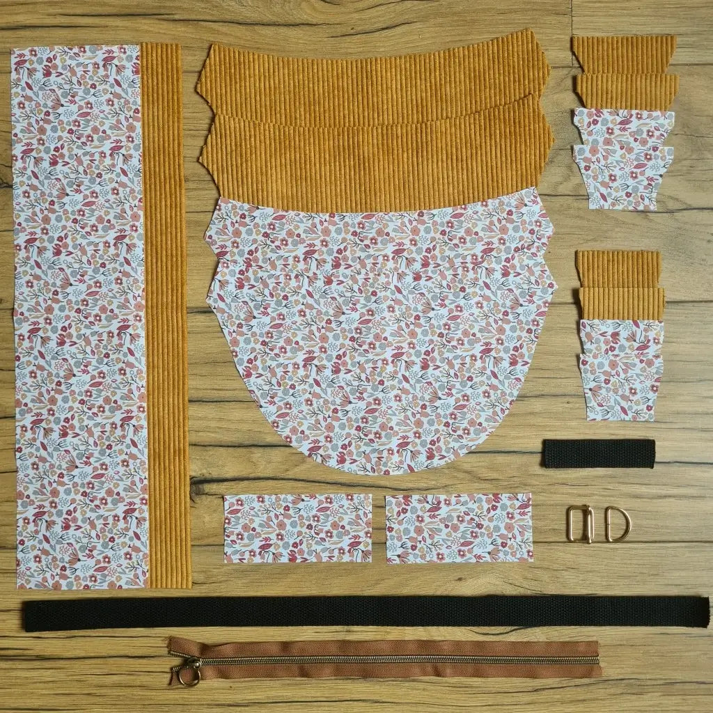 Kit "prêt à coudre" pour un sac demi-lune en velours côtelé au coloris jaune avec une doublure en tissu à motif liberty. L'ensemble des pièces de tissu, déjà prédécoupées aux bonnes dimensions, sont disposées au sol et comprennent neuf pièces en tissu à motif liberty et sept pièces en velours côtelé de couleur jaune ainsi qu'une fermeture éclair marron et une sangle noire.