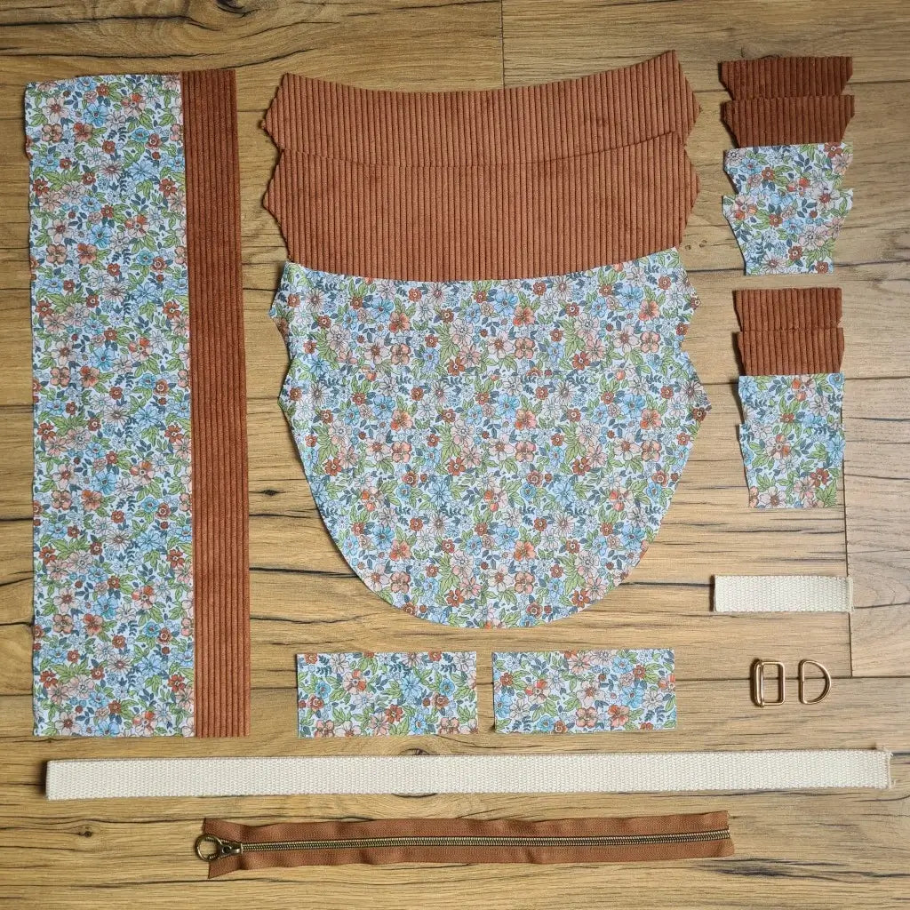 Kit "prêt à coudre" pour un sac demi-lune en velours côtelé au coloris marron avec une doublure en tissu à motif fleuri. L'ensemble des pièces de tissu, déjà prédécoupées aux bonnes dimensions, sont disposées au sol et comprennent neuf pièces en tissu à motif fleuri et sept pièces en velours côtelé de couleur marron ainsi qu'une fermeture éclair marron et une sangle beige.