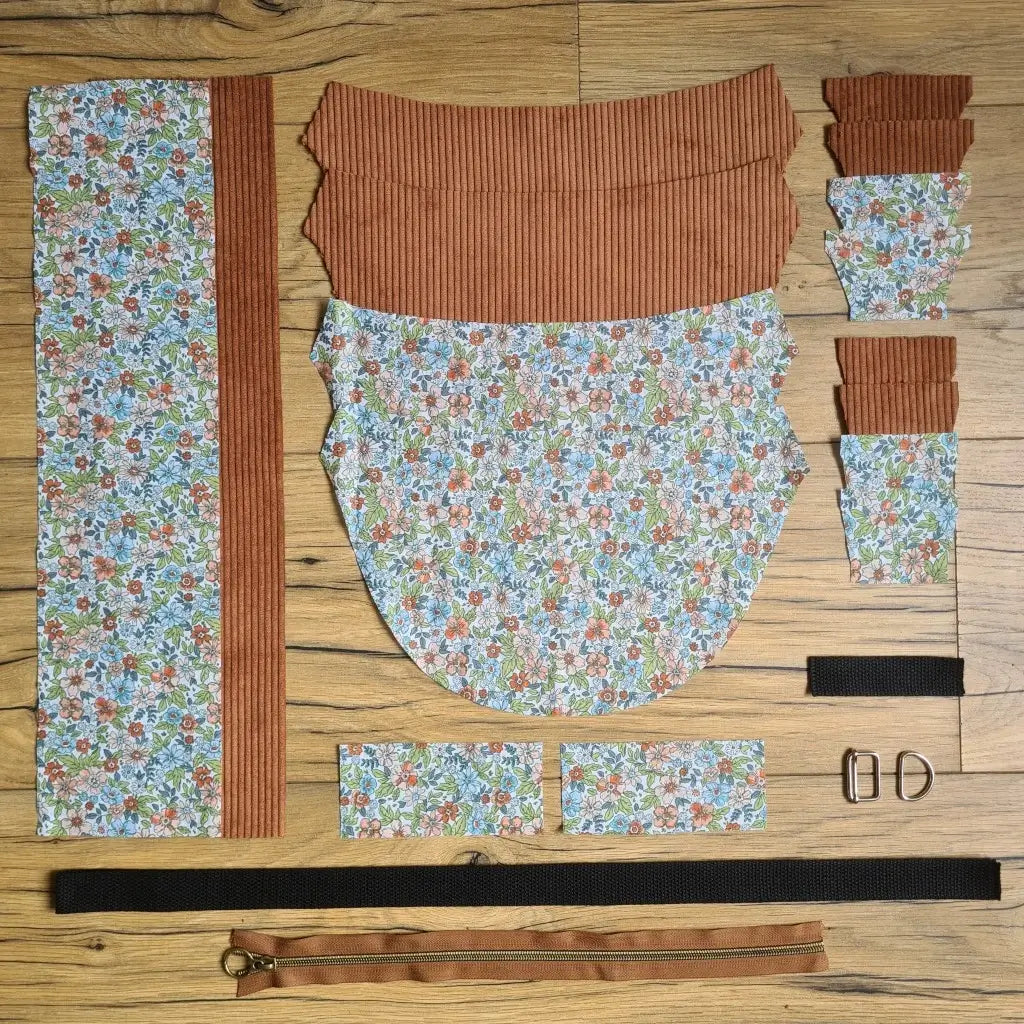 Kit "prêt à coudre" pour un sac demi-lune en velours côtelé au coloris marron avec une doublure en tissu à motif fleuri. L'ensemble des pièces de tissu, déjà prédécoupées aux bonnes dimensions, sont disposées au sol et comprennent neuf pièces en tissu à motif fleuri et sept pièces en velours côtelé de couleur marron ainsi qu'une fermeture éclair marron et une sangle noire.